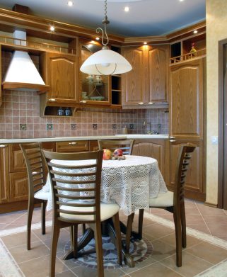 cozy kitchen: walnut cupboards, round table covered with beautiful white crocheted table-cloth with rubicund apples on it, four chairs, low pulled down lamp; pertfect illumination